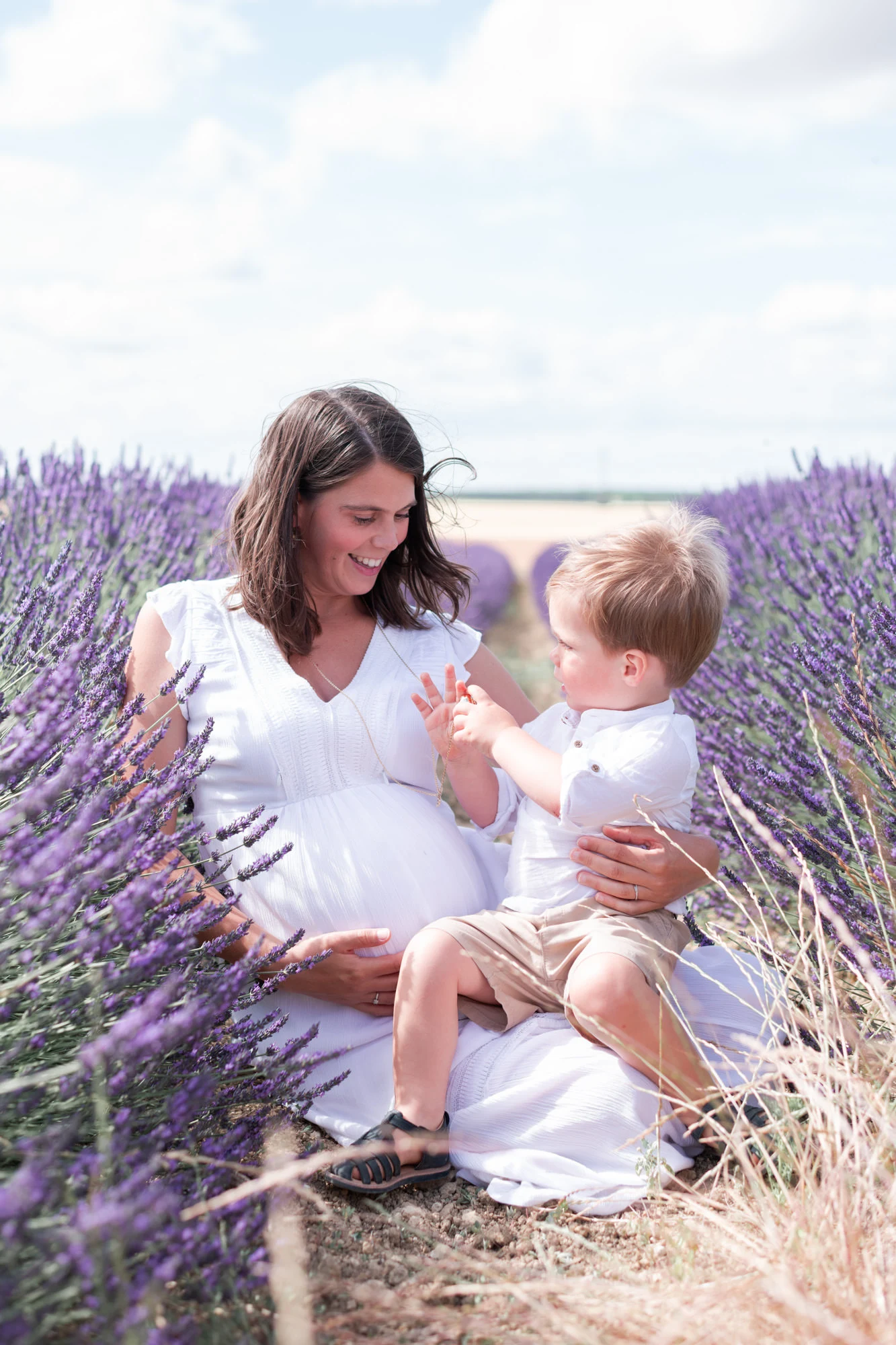 Femme enceinte et son fils aîné, habillés en blanc, lors d'une séance photo de grossesse dans les champs de lavande / lavandin fleuri en Seine et Marne