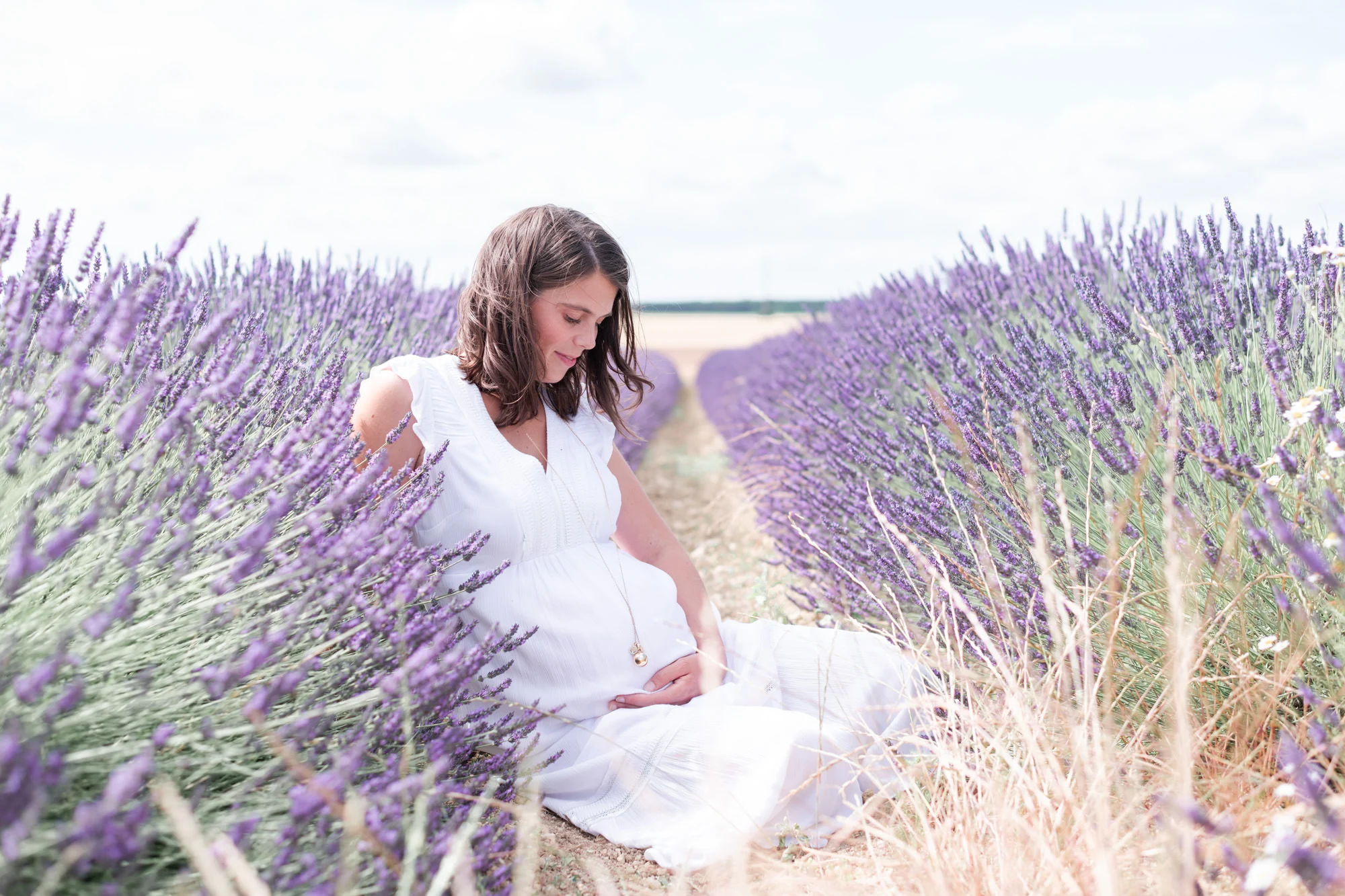 Femme enceinte, habillée en blanc, lors d'une séance photo de grossesse dans les champs de lavande / lavandin fleuri en Seine et Marne