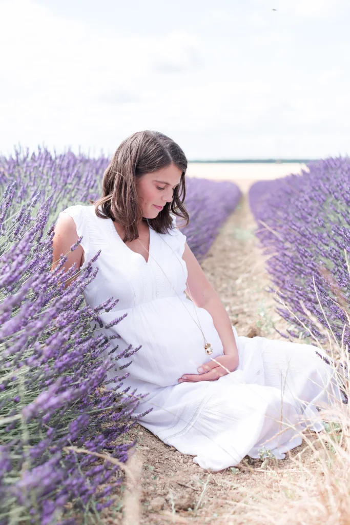 Future maman, la main sur son ventre rond, habillée en blanc et assise dans un champs de lavandin près de Fontainebleau