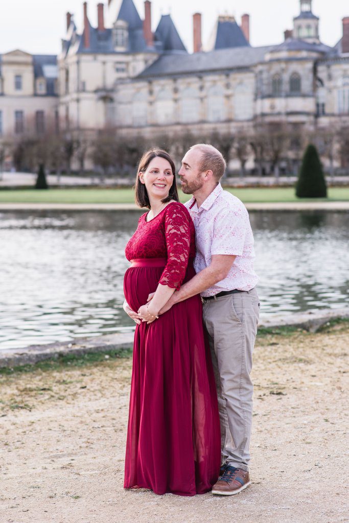 femme enceinte habillée en robe rouge bordeaux, et son mari, lors d'un shooting grossesse dans le grand parterre (jardins à la française) du château de fontainebleau