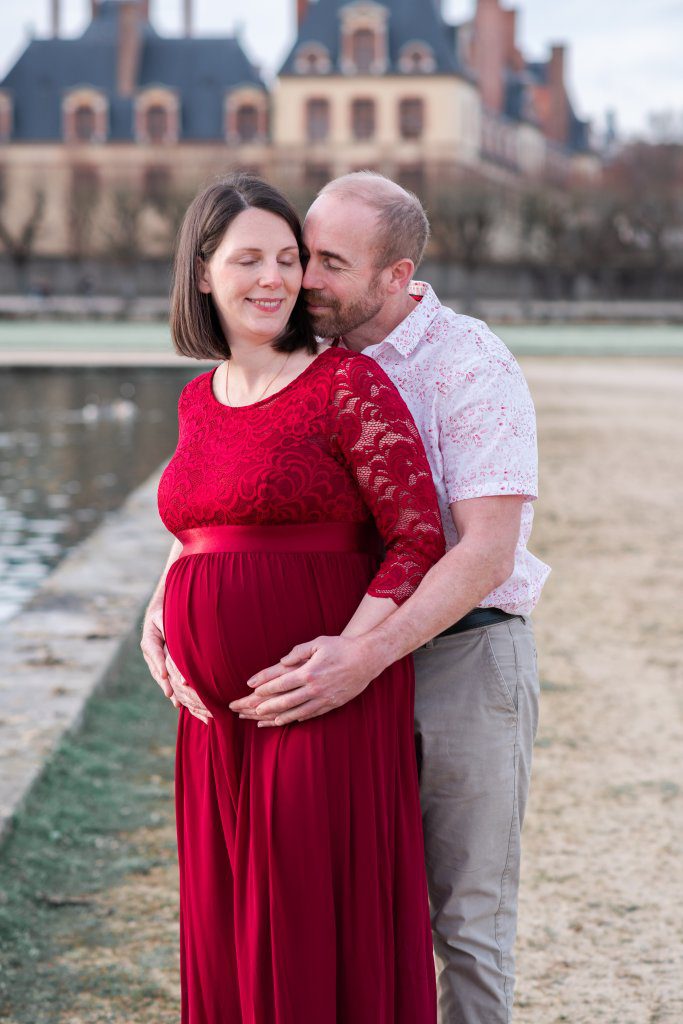 femme enceinte habillée en robe rouge bordeaux, et son mari, lors d'un shooting grossesse dans le grand parterre (jardins à la française) du château de fontainebleau