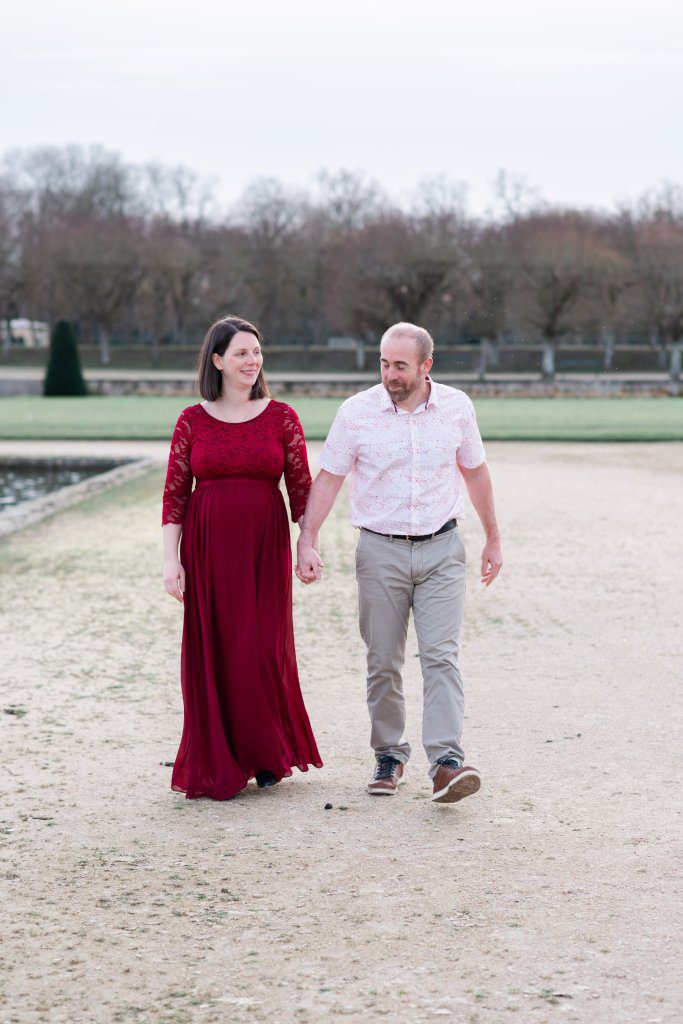 femme enceinte habillée en robe rouge bordeaux, et son mari, lors d'un shooting grossesse dans le grand parterre (jardins à la française) du château de fontainebleau