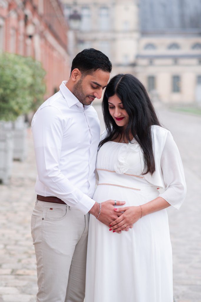 Couple habillés en blanc lors d'un shooting grossesse dans la cour d'honneur du chateau de fontainebleau avec l'escalier en fer à cheval en arrière plan